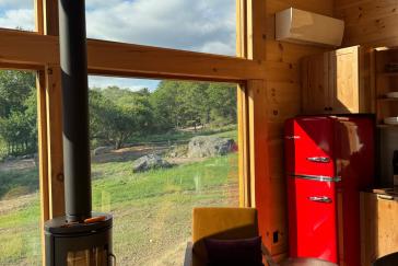 Large windows showcasing the Maine woods backdrop with a red retro refrigerator and a cozy wood burning stove.
