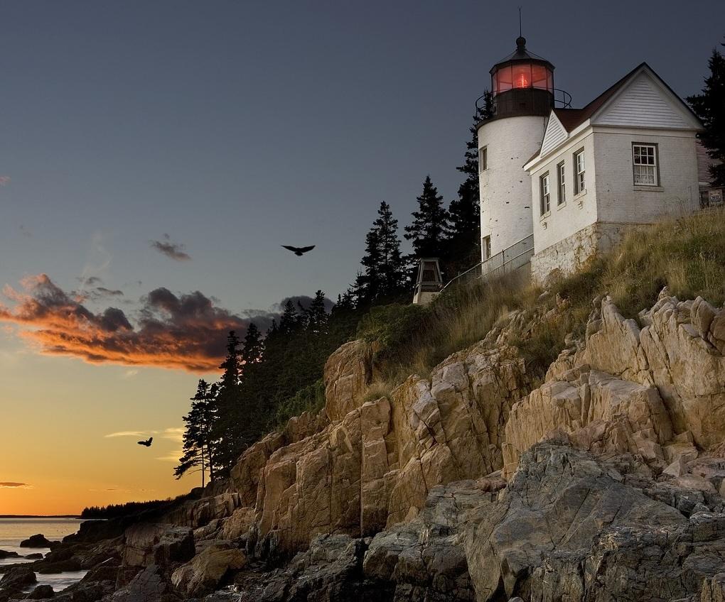 A brick lighthouse on the coast of Maine in Bar Harbor. The foundation is made of stone along a rocky cliff. The restored light glows red against a sunset.
