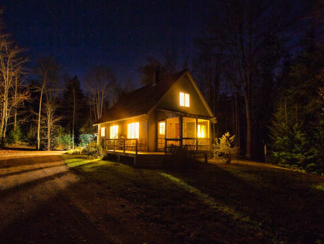 A cottage style wood sided construction at night in the woods with a starry sky and light emitting from all the windows into the surrounding woods.